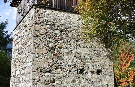 Ein Steinturm mit einem hölzernen Dach steht inmitten eines Waldes mit Herbstlaub.