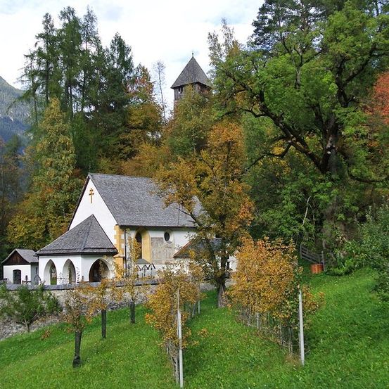 Eine kleine Kirche mit einem Turm liegt in einem üppigen grünen Tal, umgeben von Bäumen und Bergen.