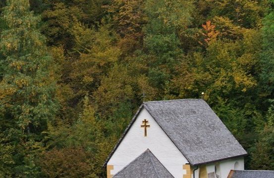 Eine kleine weiße Kirche mit einem grauen Dach steht inmitten eines dichten Waldes, mit Herbstblättern in verschiedenen Gelb- und Grüntönen. Ein Kreuz schmückt die Vorderseite des Gebäudes.