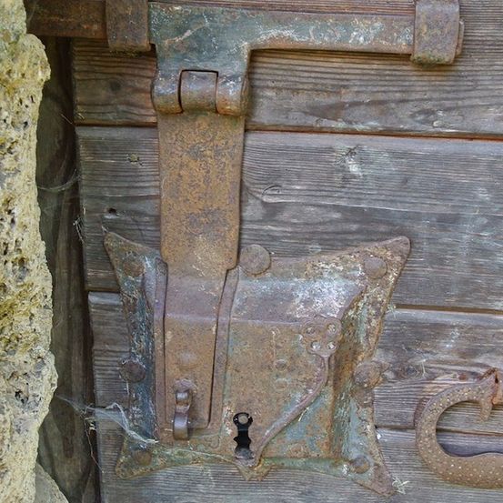 Nahaufnahme einer verrosteten Metallverriegelung an einer alten Holztür. Die Verriegelung ist mit mehreren Schrauben an der Tür befestigt und hat ein Schlüsselloch in der Mitte.