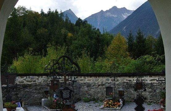 Ein ruhiger Friedhof mit Steinmauern und Blick auf die Berge, mit verschiedenen Grabsteinen und Kreuzen, umgeben von Grün.