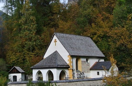 Eine kleine Kapelle mit einem grauen Dach und einem Kreuz darauf, umgeben von Bäumen und Bergen.