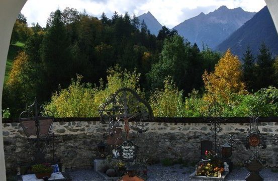 Ein ruhiger Friedhof mit Steinmauern, umgeben von üppigem Grün und entfernten Bergen unter einem klaren Himmel.