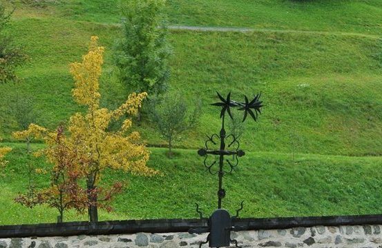 Ein Metall-Wetterhahn mit sternförmigen Spitzen steht auf einer Steinmauer und blickt auf einen grünen Hügel mit gelben und grünen Bäumen.