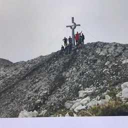 Eine Gruppe von Menschen steht auf einem felsigen Berggipfel mit einem Kreuz in der Mitte.
