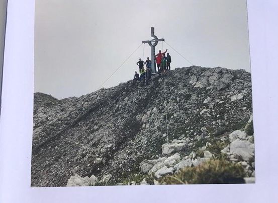 Eine Gruppe von Menschen steht auf einem felsigen Berggipfel mit einem Kreuz in der Mitte.