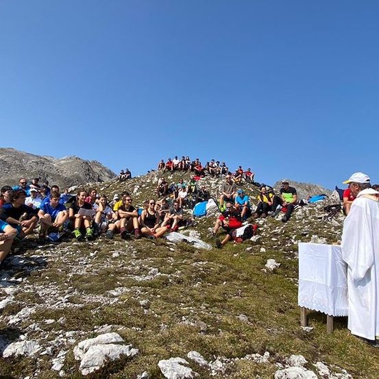 Ein Priester in einer weißen Robe steht auf einem Berg und führt eine Zeremonie mit einer Menschenmenge durch, die auf dem Boden sitzt.