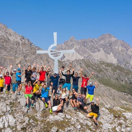 Eine Gruppe von Wanderern posiert für ein Foto auf einem Berggipfel mit einem großen Kreuz im Hintergrund.