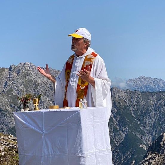 Ein Priester steht an einem Altar auf einem Berggipfel, umgeben von Blumen und einem goldenen Kelch. Die Umgebung ist ruhig mit Bergen im Hintergrund.