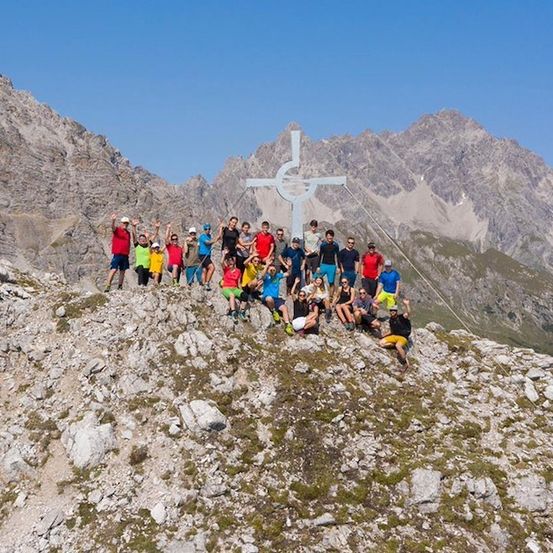 Eine Gruppe von Wanderern posiert für ein Foto auf einem Berggipfel mit einem großen Kreuz im Hintergrund.