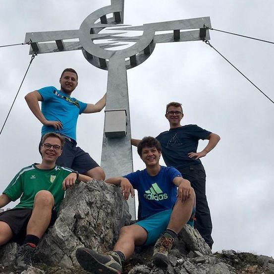 Vier Männer posieren für ein Foto auf einem Berggipfel, mit einer großen Kreuzstruktur im Hintergrund.