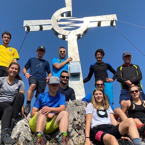 Eine Gruppe von Wanderern posiert für ein Foto auf einem Berggipfel mit einem großen Kreuz im Hintergrund.