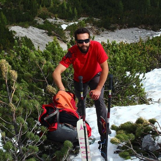 Ein Mann in einem roten Shirt und Sonnenbrille steht auf einem verschneiten Berg und hält Skistöcke und einen Rucksack.