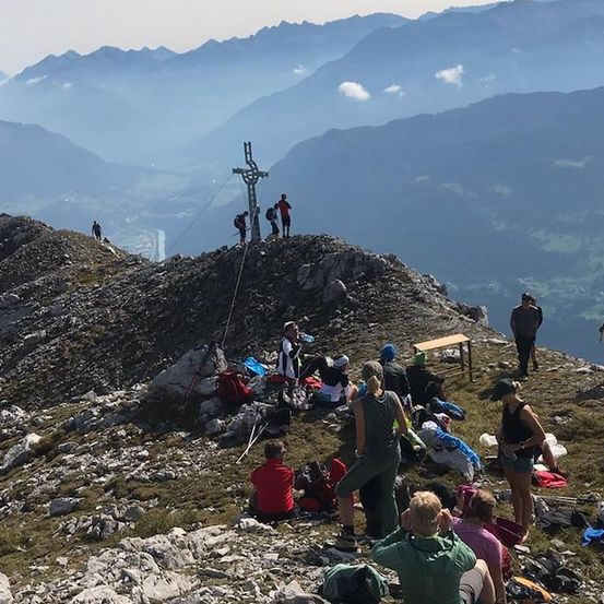 Eine Gruppe von Wanderern hat sich auf dem Gipfel eines Berges versammelt, wo ein Kreuz den Gipfel markiert. Einige sitzen, andere stehen, und um sie herum sind Rucksäcke und Ausrüstung verstreut.