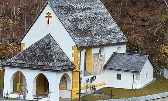 Eine kleine weiße Kirche mit grauem Dach und gelben Akzenten, mit einem Kreuz auf der Spitze und einem kleinen Nebengebäude. Sie ist von einer Steinmauer und saftigem grünem Gras umgeben.