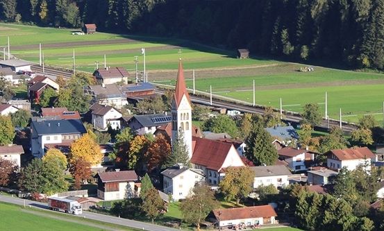 Ein Luftbild eines kleinen Dorfes mit einer Kirche, umgeben von grünen Feldern und Bäumen, mit einer Straße und einer Eisenbahn in der Ferne.