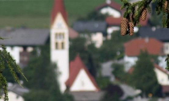 Ein Dorf mit einem Kirchturm und roten Dächern, umgeben von Bäumen und Grün. Das Bild ist leicht verschwommen.