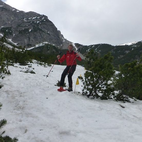 Eine Person in einem roten Jackett treibt Langlauf auf einem verschneiten Bergweg mit Kiefern und Bergen im Hintergrund.