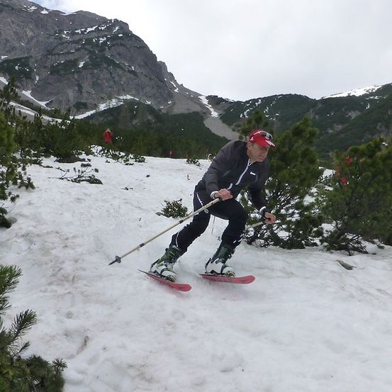 Ein Mann skilangt einen schneebedeckten Berg hinunter und hält Skistöcke. Er trägt eine rote Mütze und eine schwarze Jacke.