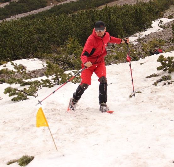 Ein Mann in einem roten Schneeanzug schippert einen schneebedeckten Hügel hinauf und hält Skistöcke.
