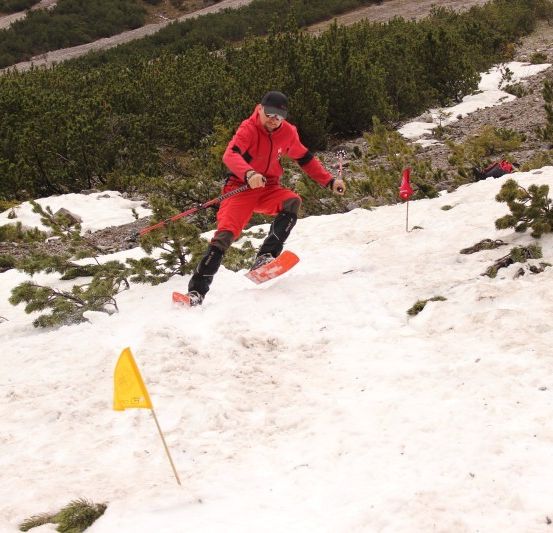 Ein Skifahrer in einem roten Schneeanzug navigiert einen verschneiten Hang und hält Skistöcke, mit einer gelben Flagge, die den Weg markiert.