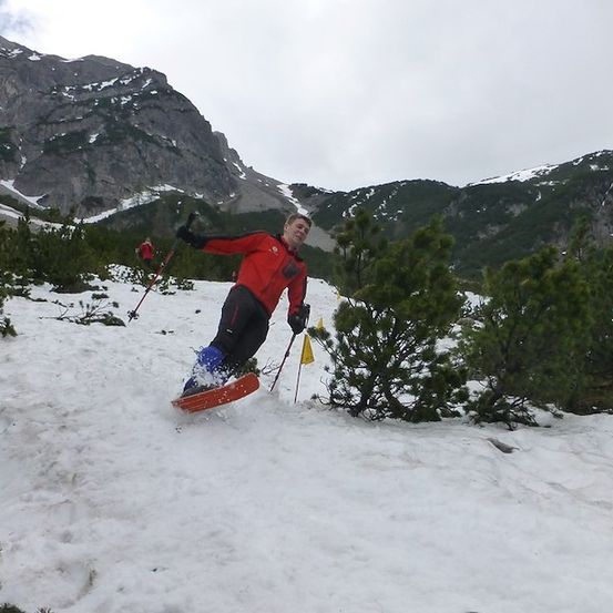 Ein Mann in einem roten Jacken fährt mit Skistöcken bergab auf einem verschneiten Berg.