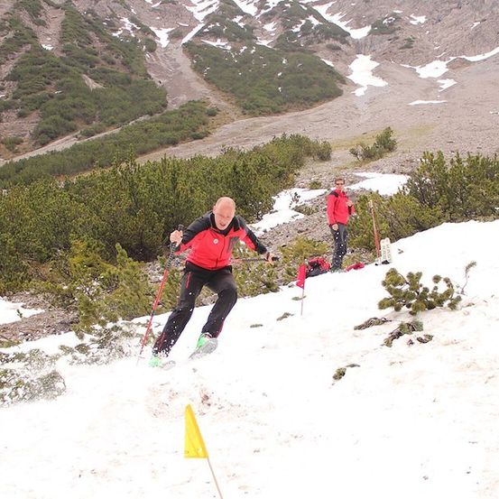 Ein Mann in einem roten Jackett skilangs einen schneebedeckten Berg hinunter, mit einer weiteren Person hinter ihm.
