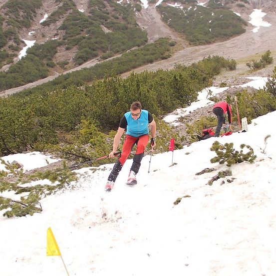 Ein Skifahrer navigiert einen schneebedeckten Berghang und benutzt Skistöcke zum Ausgleich. Ein weiterer Skifahrer ist dahinter und justiert etwas am Boden.