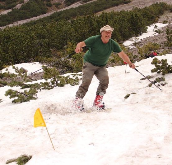 Ein Mann in einem grünen Shirt und Hut skifahrer einen schneebedeckten Hang hinunter, hält Skistöcke und hat eine gelbe Fahne im Schnee.