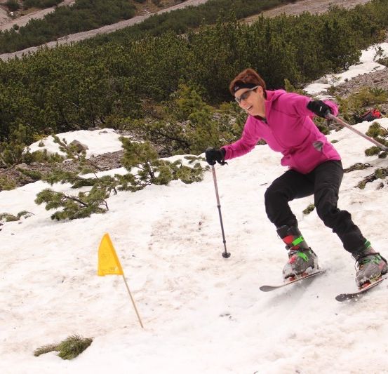 Eine Frau in einer pinken Jacke skifahrert einen schneebedeckten Hang hinunter, hält Skistöcke und trägt eine Sonnenbrille. Eine gelbe Fahne markiert ihren Weg.