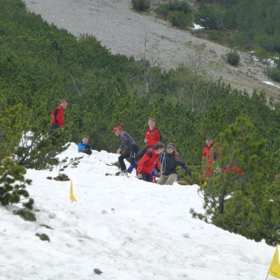 Eine Gruppe von Menschen in roten Jacken und Mützen skifahren einen schneebedeckten Berg hinunter, wobei einige gelbe Flaggen den Weg markieren.