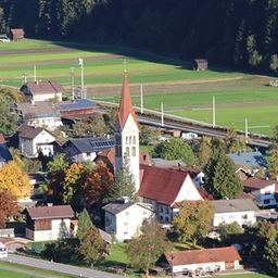 Luftaufnahme eines kleinen Dorfes mit einer Kirche, umgeben von grünen Feldern und Bäumen. Das Dorf liegt zwischen einer Straße und einer Eisenbahnstrecke.