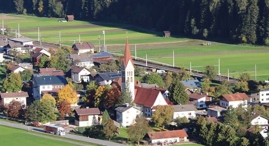 Luftaufnahme eines kleinen Dorfes mit einer Kirche, umgeben von grünen Feldern und Bäumen. Das Dorf liegt zwischen einer Straße und einer Eisenbahnstrecke.