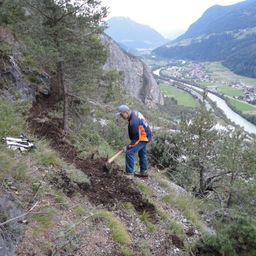 Ein Mann in einem orangefarbenen und blauen Jackett gräbt an einem steilen Hang mit einem Fluss und Bergen im Hintergrund.