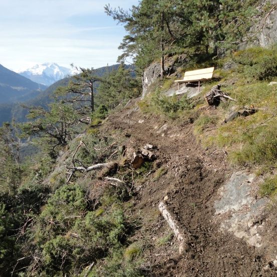 Eine Holzbank an einem steilen Hang mit Blick auf Berge und Bäume.