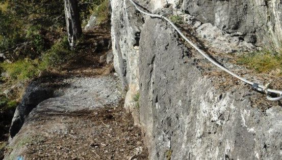 Ein felsiger Abhang mit einem befestigten Metallseil, möglicherweise zum Klettern. Der Weg ist schmal und von Grün umgeben.