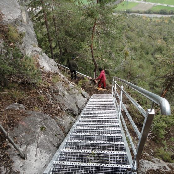 Eine Metalltreppe mit Handlauf ist an einem steilen Berghang errichtet. Zwei Personen befinden sich am Hang, eine in einer roten Jacke und die andere in einer schwarzen Jacke.