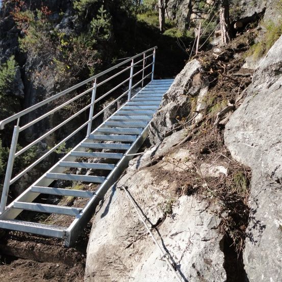 Eine Metalltreppe mit Handläufen ist an einem felsigen Berghang befestigt, umgeben von Grün.