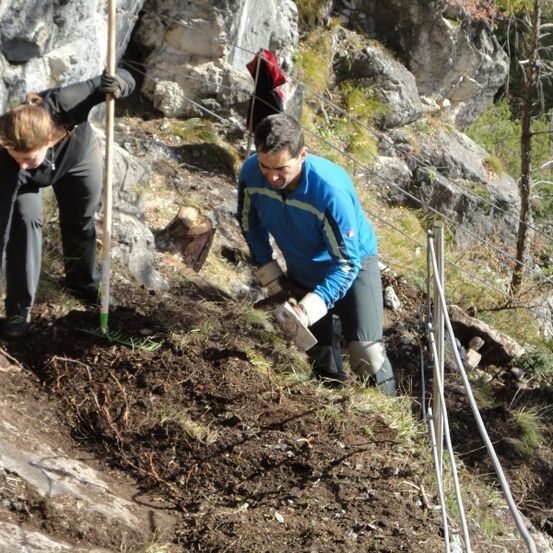 Zwei Personen graben an einem steilen Hang, einer trägt einen schwarzen und der andere einen blauen Mantel. Sie benutzen Werkzeuge und im Hintergrund sind Felsen und Pflanzen zu sehen.
