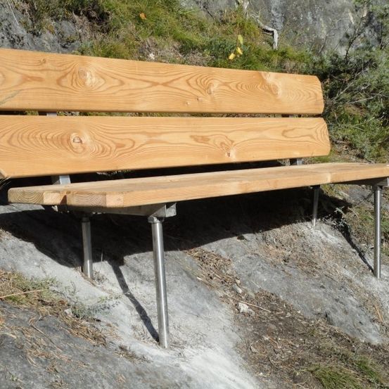 Eine Holzbank mit Metallrahmen steht auf einer felsigen Oberfläche. Die Bank hat ein einfaches Design mit einer flachen Rückenlehne und einer Sitzfläche. Die Metallbeine sind stabil und unterstützen die Bank fest.