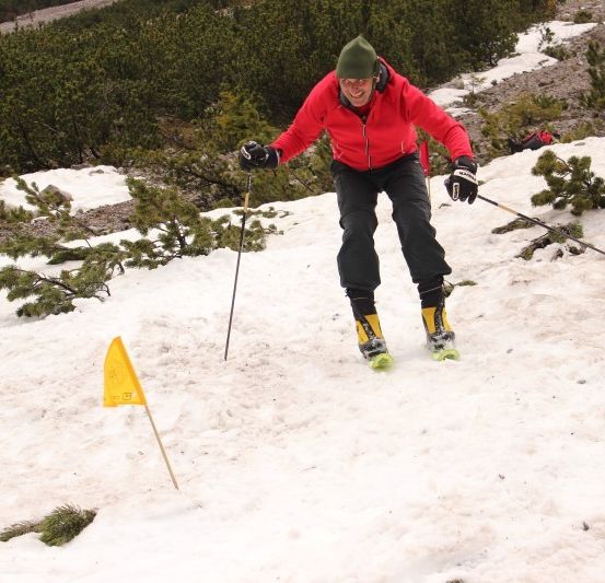 Ein Skifahrer in einem roten Jackett und schwarzen Hosen fährt einen verschneiten Hang hinunter und hält Skistöcke.