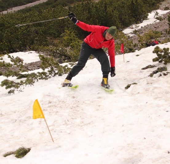 Ein Skifahrer in einem roten Jackett und grünem Hut fährt einen Schneehang hinunter und hält einen Skistock. Ein gelber Flaggenpfosten ist im Schnee.