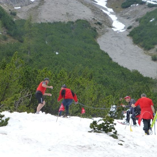 Eine Gruppe von Menschen in roten Kleidungsstücken skilt einen schneebedeckten Berg hinauf. Sie halten Skistöcke und einige machen Fotos.