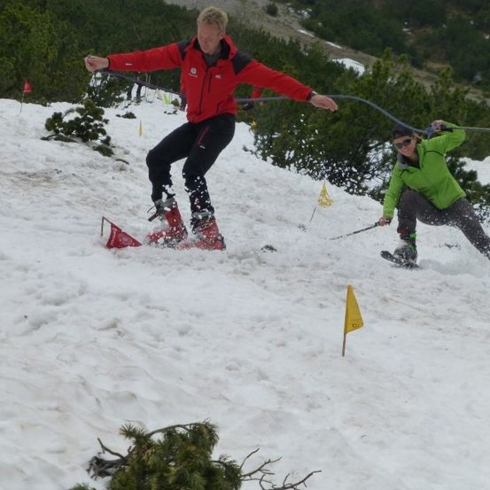 Zwei Skifahrer navigieren einen verschneiten Hang, einer in einem roten Jackett und der andere in Grün, beide halten Skistöcke.