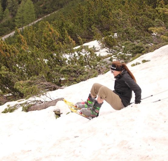 Eine Frau sitzt auf einem verschneiten Hang, trägt Skier und hält Skistöcke. Sie ist von Kiefern umgeben und einem gelben Flaggen.