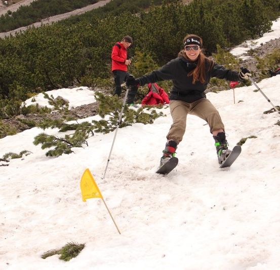 Eine Frau in einer schwarzen Jacke und Sonnenbrille fährt mit Skistöcken auf einem verschneiten Hang Ski. Ein Mann in einer roten Jacke ist im Hintergrund.
