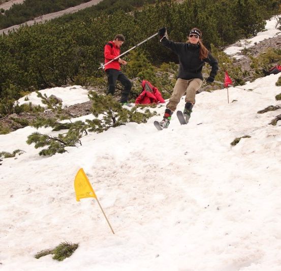 Ein Skifahrer in einer schwarzen Jacke und mit Skibrille fährt einen schneebedeckten Berg hinunter, mit einer gelben Fahne im Vordergrund. Ein weiterer Skifahrer in einer roten Jacke folgt dahinter.