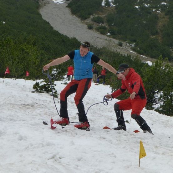 Eine Gruppe von Menschen skifahren auf einem verschneiten Berg. Eine Person hält eine Seil und Skistöcke, während eine andere Person das Seil hält.