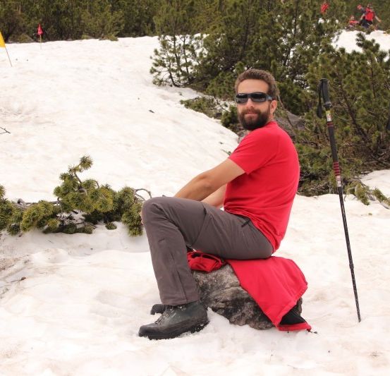 Ein Mann in einem roten Shirt und Sonnenbrille sitzt auf einem Felsen im Schnee, umgeben von Kiefern und Skistöcken.