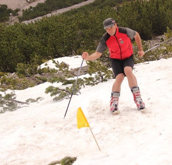 Ein Mann skis auf einem verschneiten Hügel, trägt eine rote Weste und schwarze Shorts. Er hält Skistöcke und passiert eine gelbe Flagge.